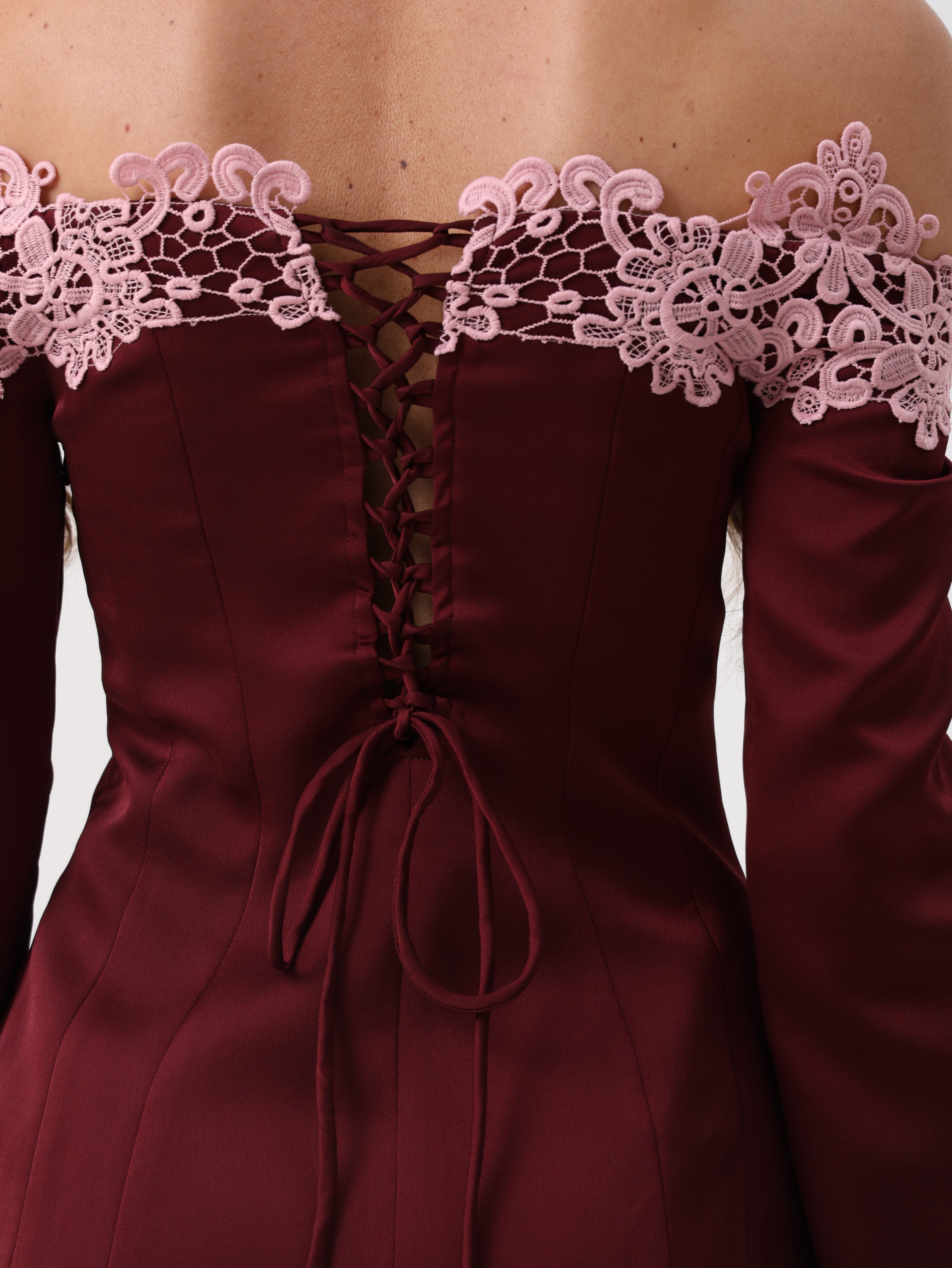 Close-up of the back of an off-the-shoulder burgundy dress with long sleeves, a lace-up corset-style back, and light pink floral lace trim across the shoulders.