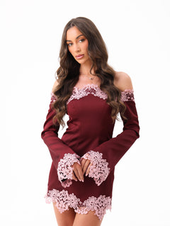 A woman modeling a burgundy off-the-shoulder long-sleeve mini dress with light pink floral lace trim at the neckline, cuffs, and hem, set against a plain white background.