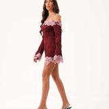 A woman modeling a burgundy off-the-shoulder mini dress with light pink floral lace trim at the neckline, cuffs, and hem, paired with clear high-heeled mules, against a plain white studio background.