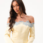 A woman modeling an off-the-shoulder pale yellow dress with light blue lace trim across the neckline, set against a plain white background.