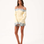 A woman modeling a pale yellow off-the-shoulder mini dress with light blue lace trim, paired with clear high-heeled sandals, against a plain white studio background.