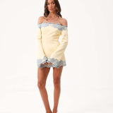 A woman modeling a pale yellow off-the-shoulder mini dress with light blue lace trim, paired with clear high-heeled sandals, against a plain white studio background.