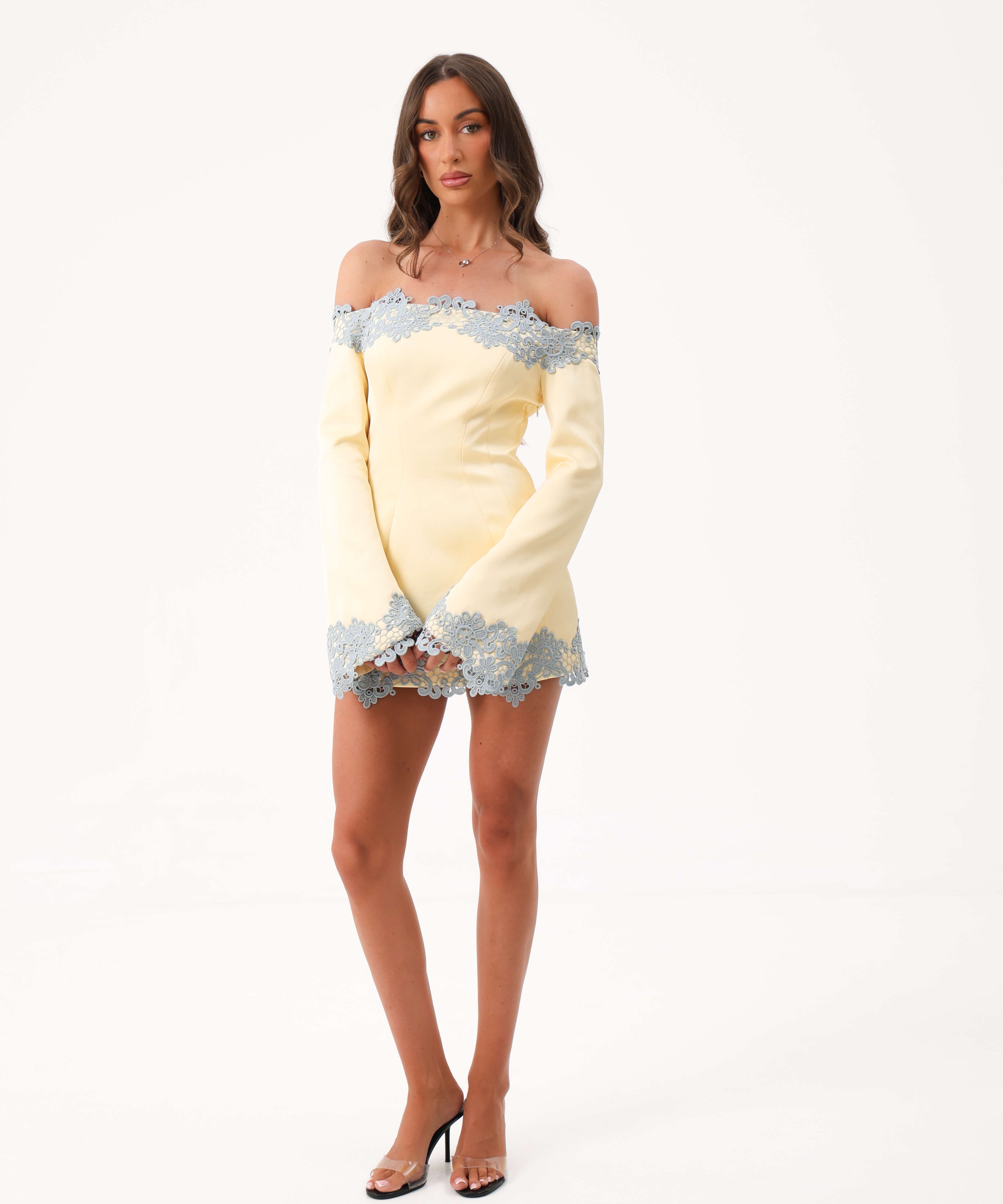 A woman modeling a pale yellow off-the-shoulder mini dress with light blue lace trim, paired with clear high-heeled sandals, against a plain white studio background.