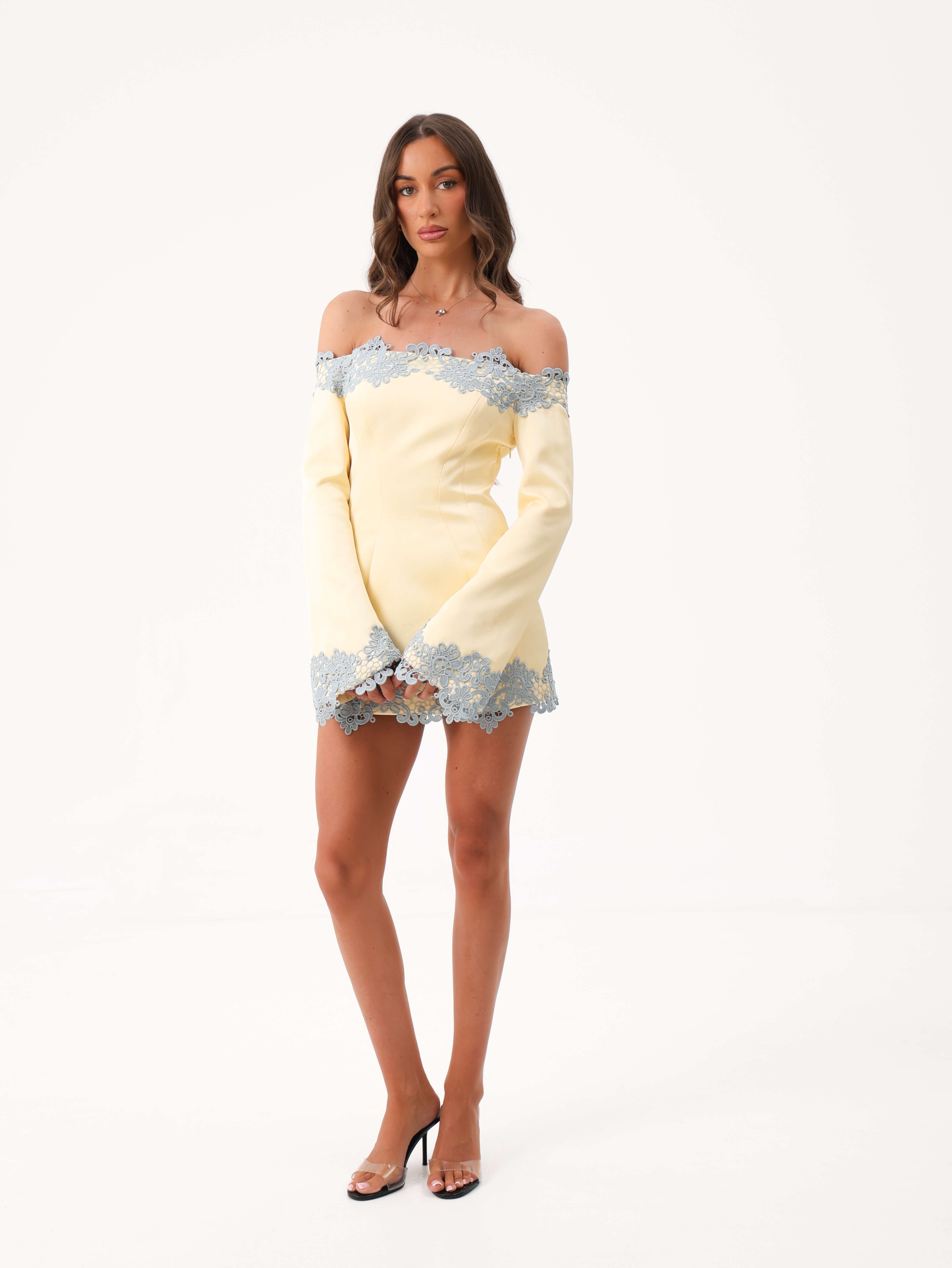 A woman modeling a pale yellow off-the-shoulder mini dress with light blue lace trim, paired with clear high-heeled sandals, against a plain white studio background.