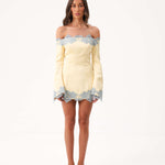 A woman modeling a yellow off the shoulder mini dress with blue lace detailing against a white studio background.