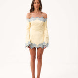 A woman modeling a yellow off the shoulder mini dress with blue lace detailing against a white studio background.
