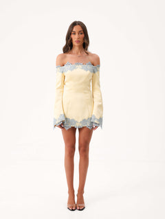 A woman modeling a yellow off the shoulder mini dress with blue lace detailing against a white studio background.