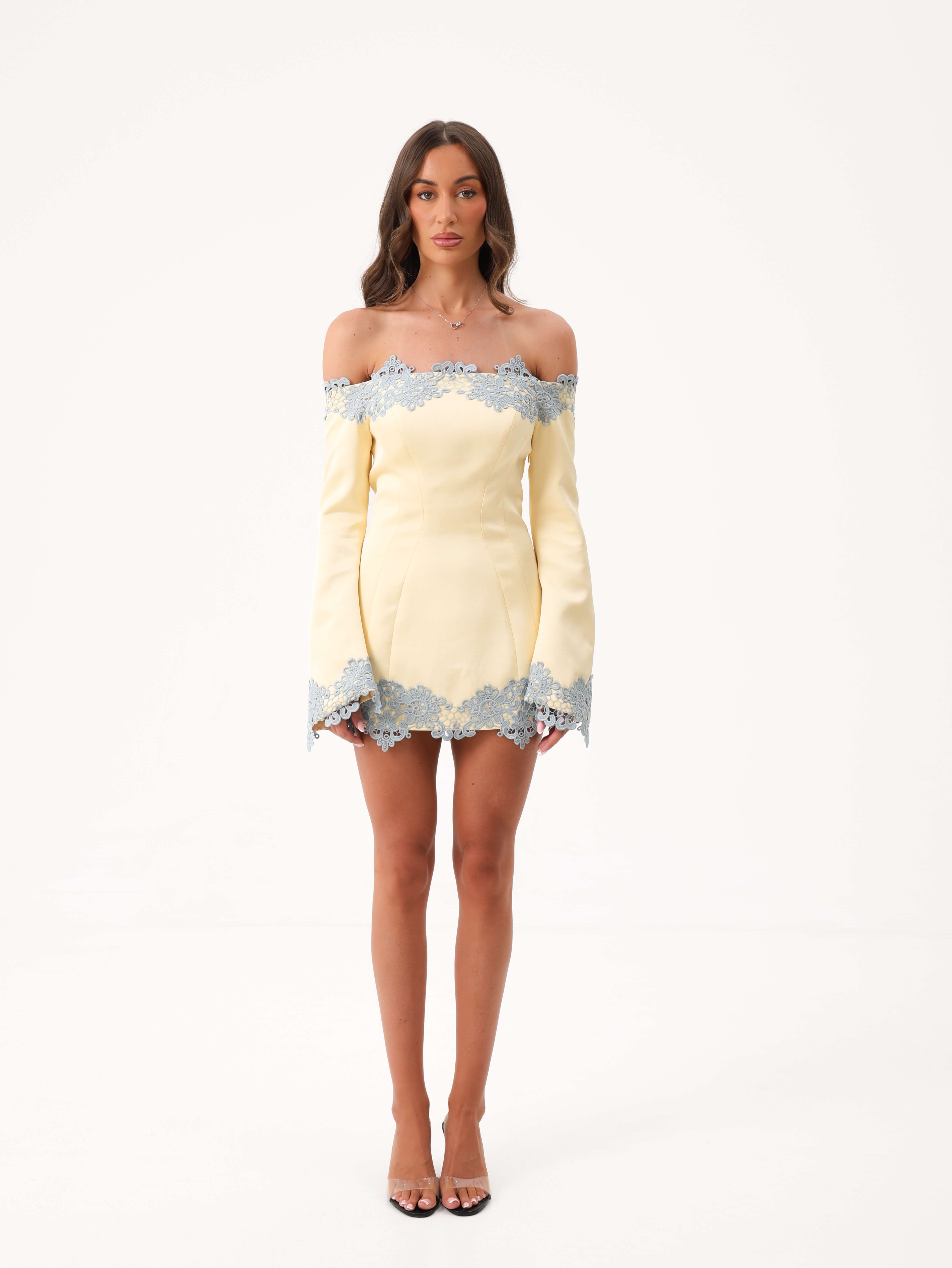 A woman modeling a yellow off the shoulder mini dress with blue lace detailing against a white studio background.