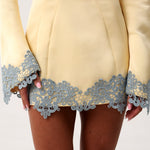 Close-up of a pale yellow dress with long sleeves and a decorative light blue lace trim along the hem and cuffs, worn by a model.