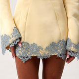 Close-up of a pale yellow dress with long sleeves and a decorative light blue lace trim along the hem and cuffs, worn by a model.