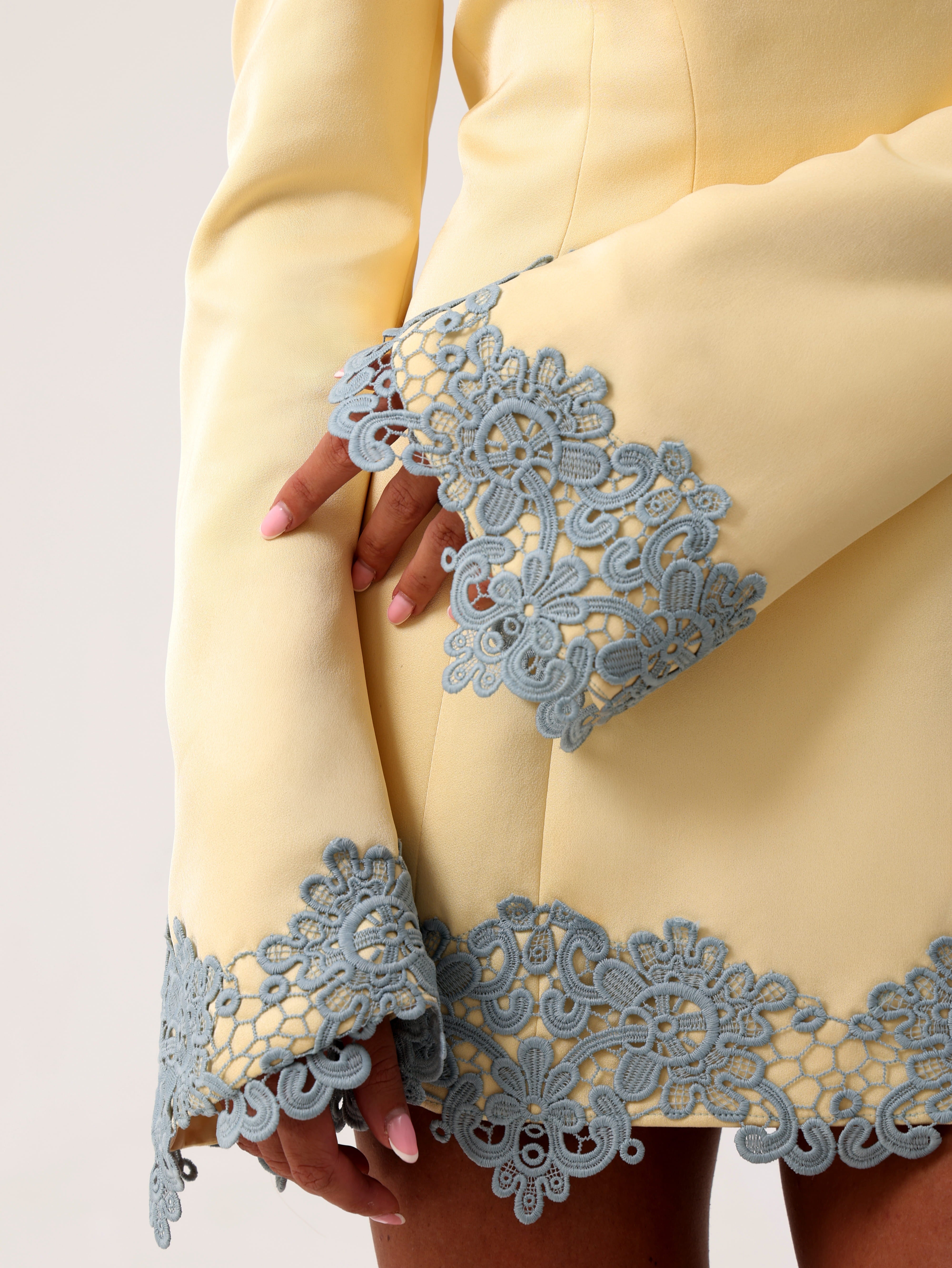 Close-up of a pale yellow dress with long flared sleeves and dusty blue lace trim at the cuffs and hem, shown on a model.